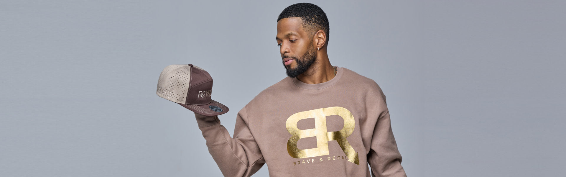 A person wearing a beige sweatshirt with a gold logo holds a brown and beige cap, set against a muted background.