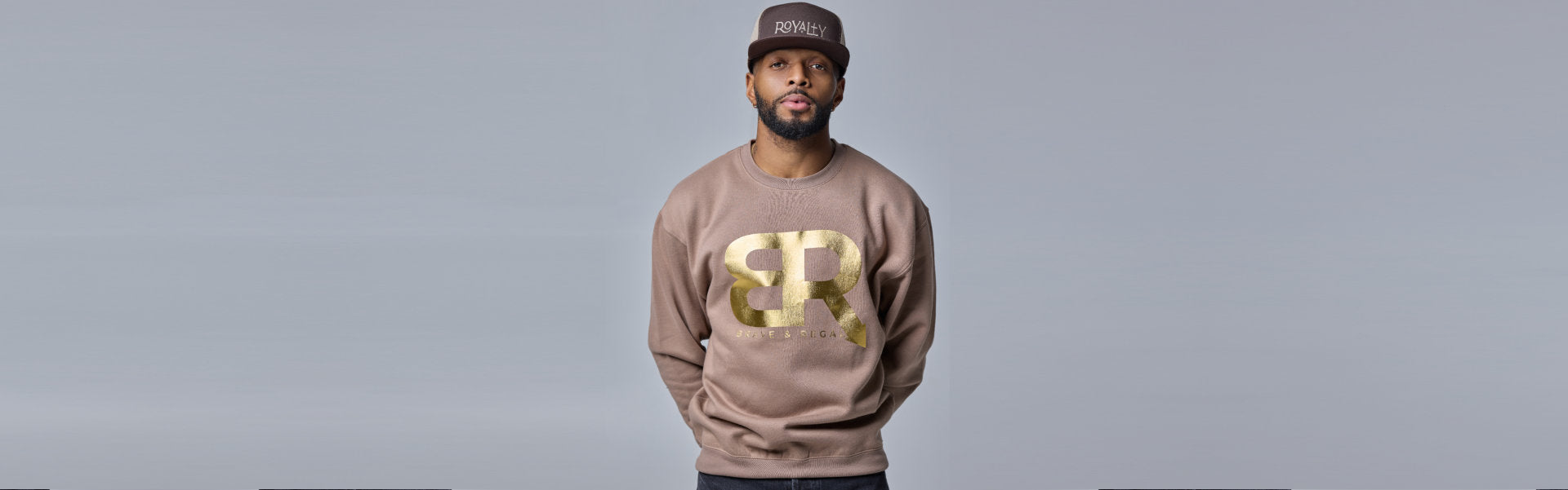 Person wearing a beige sweatshirt with a large gold BR logo and text BALENCIAGA underneath, paired with a dark cap.