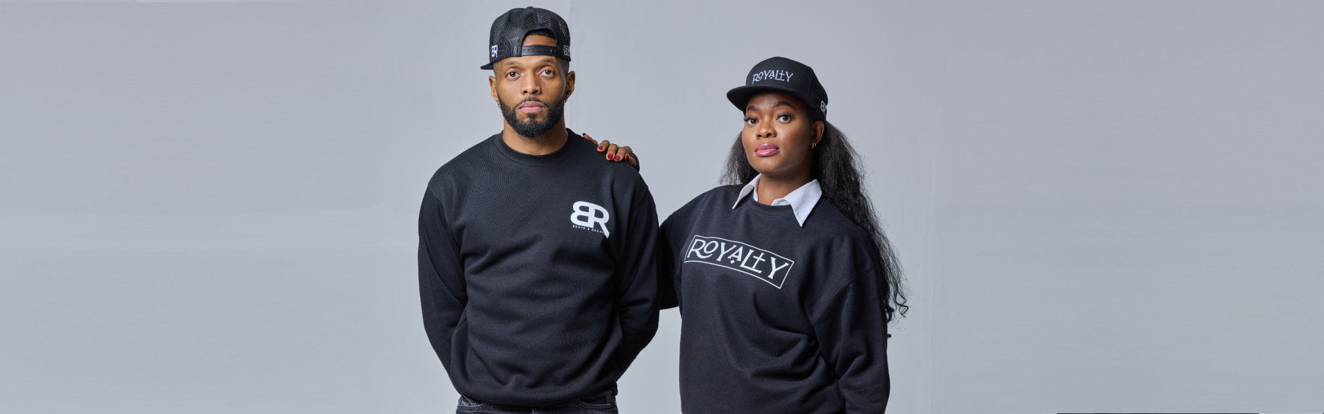 Two individuals stand side by side, wearing matching black sweatshirts with white logos, against a gray background.