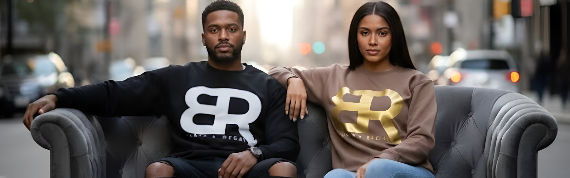 Two individuals sit on a gray couch in an urban setting, wearing matching sweatshirts with bold BR logos.