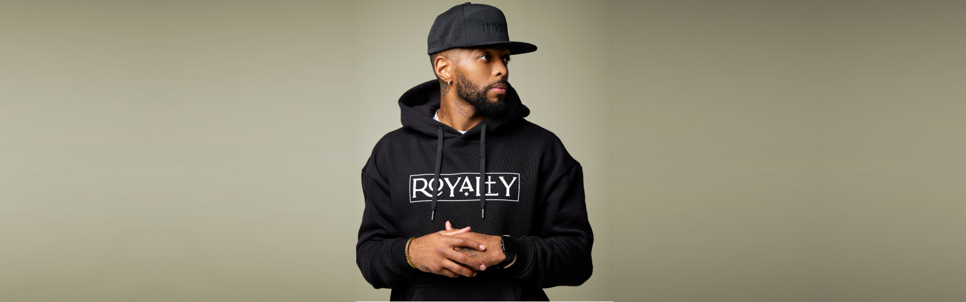Person wearing a black hoodie with ROYALTY printed on it, a black cap, and a watch, standing against a beige background.