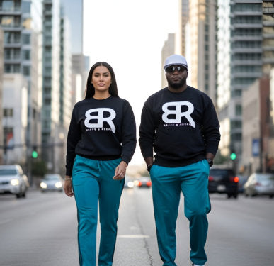 Two individuals wearing matching black sweatshirts and teal pants walk confidently down a city street.